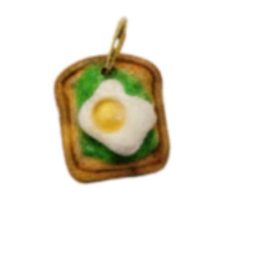 A small food-themed charm or pendant featuring a slice of toast with a fried egg and green leaves, centered on a plain white background.