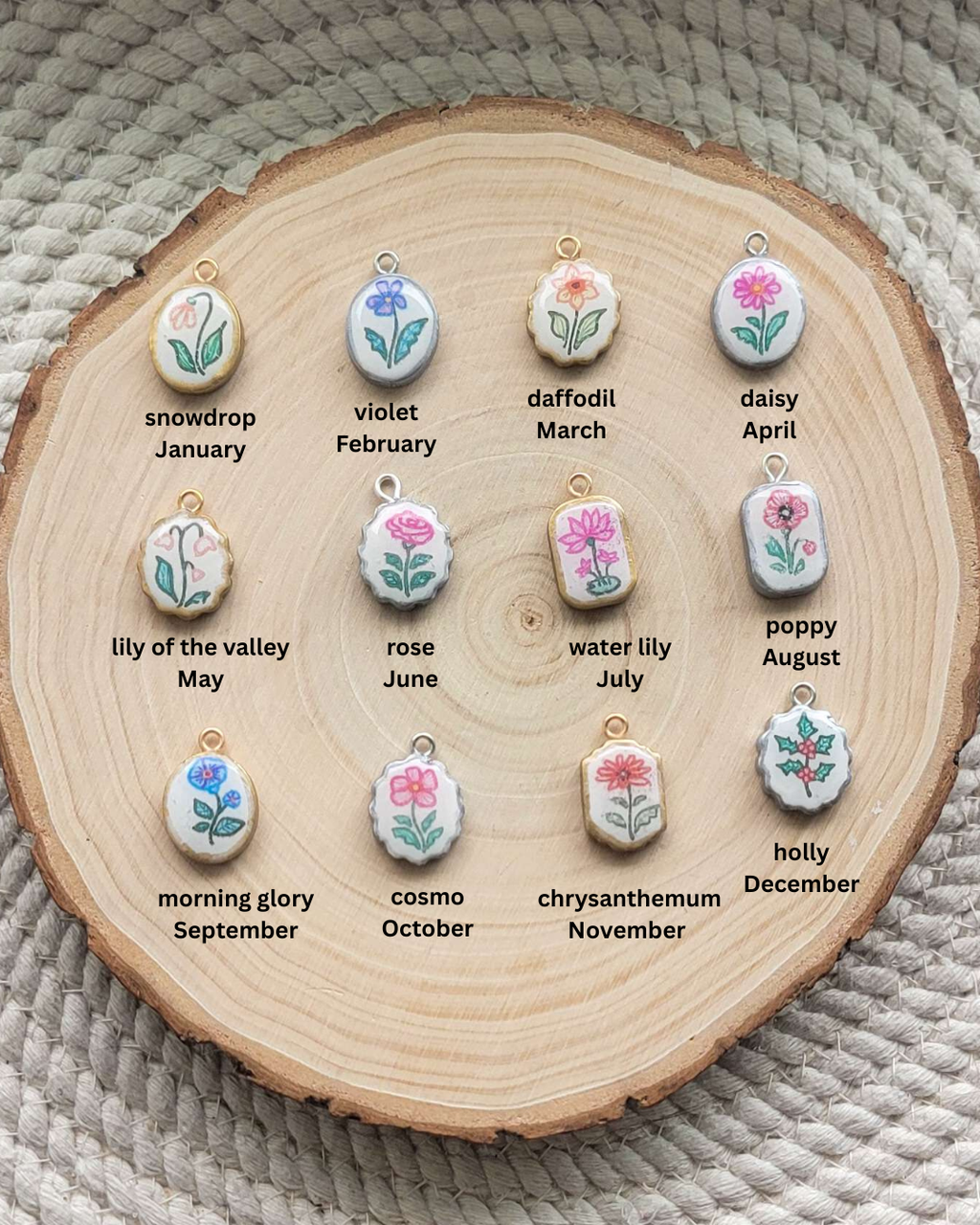 Twelve small floral pendant charms arranged on a round wood slice, each labeled with a flower name and month. The pendants are white with painted flowers in pink, blue, green, and yellow, and small gold-tone loops at the top.