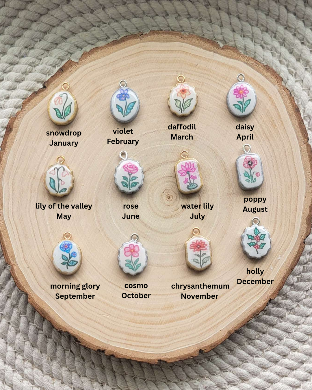 Twelve small floral pendant charms arranged on a round wood slice, each labeled with a flower name and month. The pendants are white with painted flowers in pink, blue, green, and yellow, and small gold-tone loops at the top.