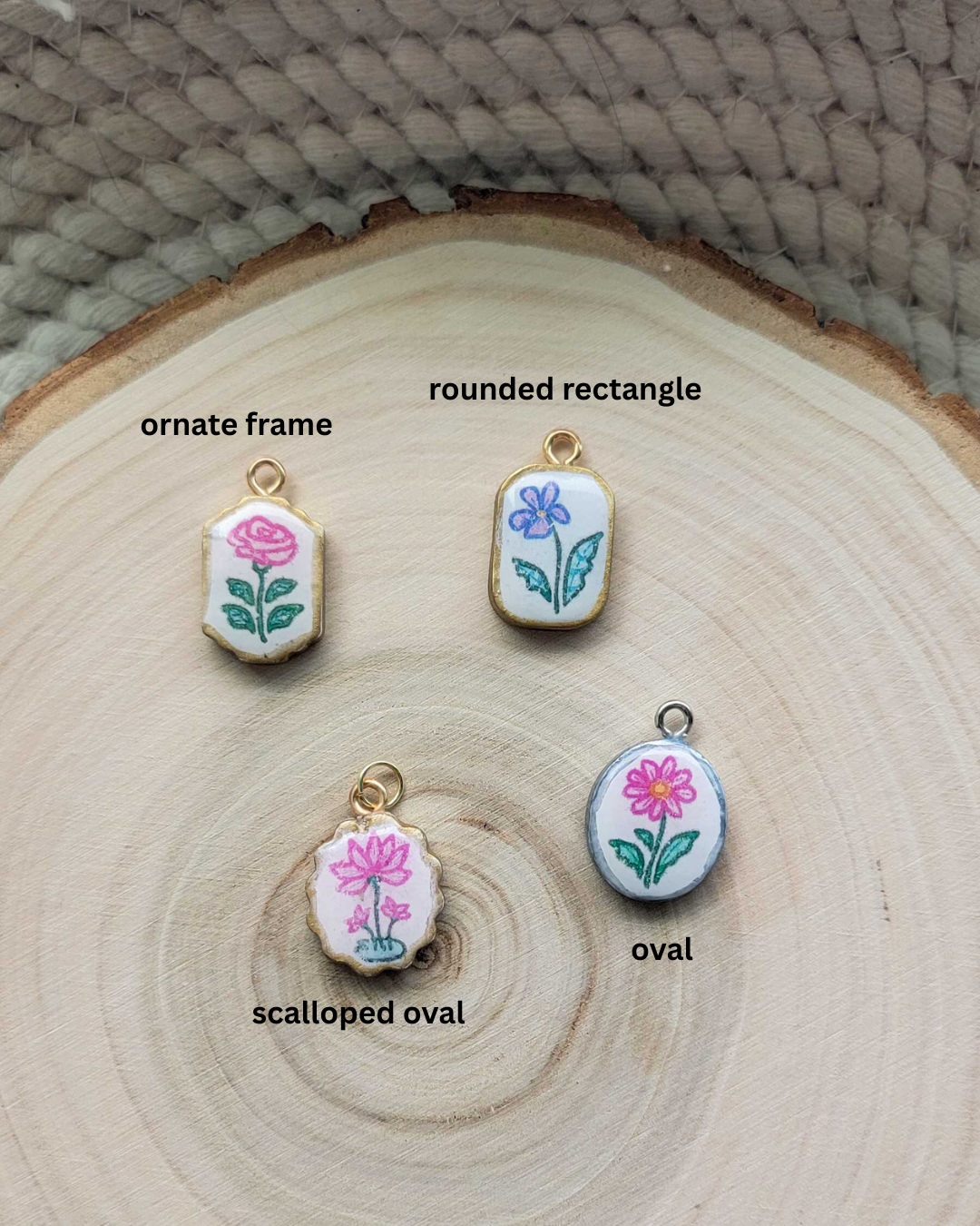Four small floral enamel-style charms or pendants displayed on a round wood slice background, labeled 'ornate frame,' 'rounded rectangle,' 'scalloped oval,' and 'oval.' The charms have white faces with pink flowers, green leaves, and gold-tone edges, and are arranged near a woven neutral surface.