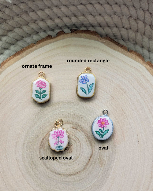 Four small floral enamel-style charms or pendants displayed on a round wood slice background, labeled 'ornate frame,' 'rounded rectangle,' 'scalloped oval,' and 'oval.' The charms have white faces with pink flowers, green leaves, and gold-tone edges, and are arranged near a woven neutral surface.