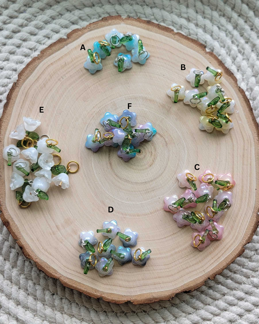Six clusters of small flower-shaped beads labeled A through F, arranged on a round wood slice over a woven beige textile background. The beads come in pastel blue, lavender, pink, and white with green and gold accents.
