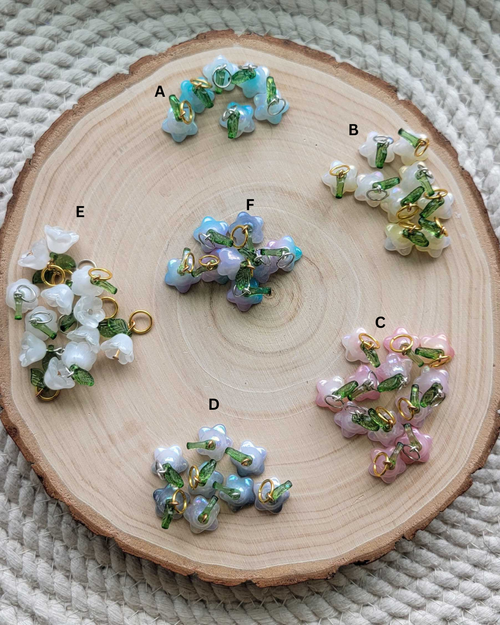 Six clusters of small flower-shaped beads labeled A through F, arranged on a round wood slice over a woven beige textile background. The beads come in pastel blue, lavender, pink, and white with green and gold accents.
